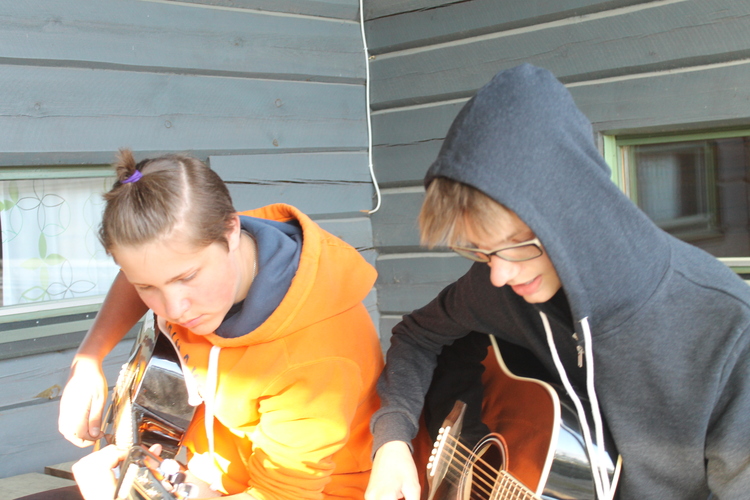Två ungdomar spelar gitarr utanför en lägergård.
